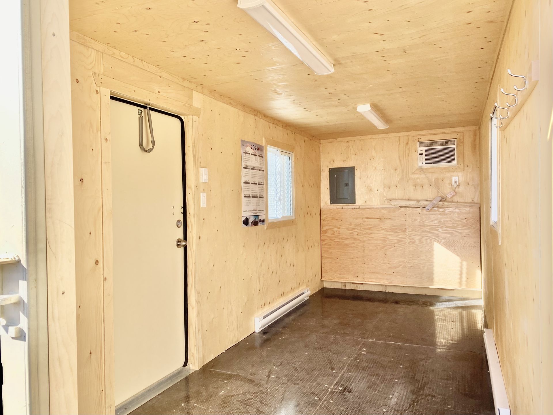 Intérieur d’un conteneur bureau de chantier avec murs en contreplaqué, éclairage au plafond, fenêtre, panneau électrique et climatiseur mural.