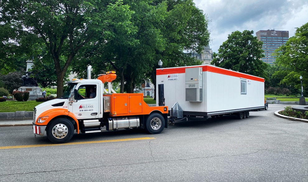 Camion transportant une roulotte de chantier blanche et orange vers un site de construction temporaire.